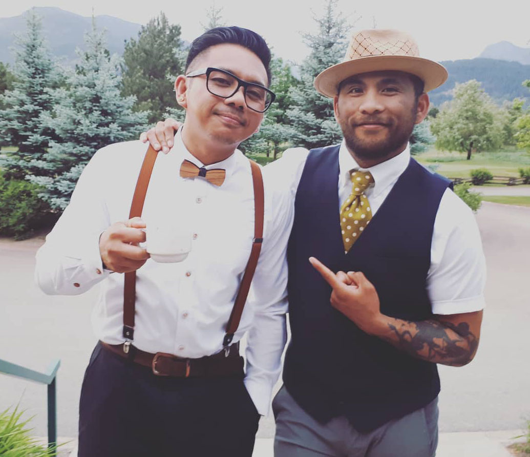 Two well-dressed men, Marc and Stew, standing outdoors and smiling while enjoying a light moment in formal attire.